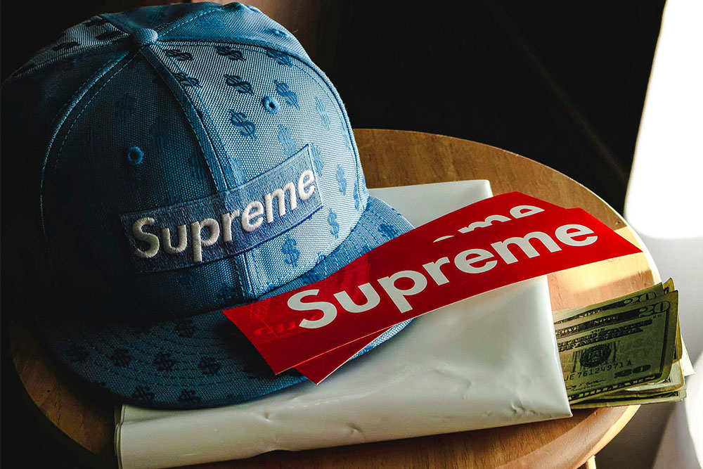 brand supreme with red supreme stickers and blue supreme hat on table with money in a bag