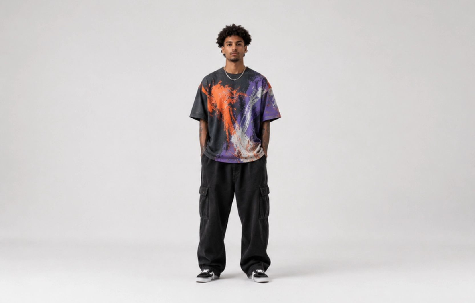 Young man stands facing camera in an oversized multicolor splatter-painted T-shirt with black cargo pants on a neutral studio backdrop, sneakers visible.
