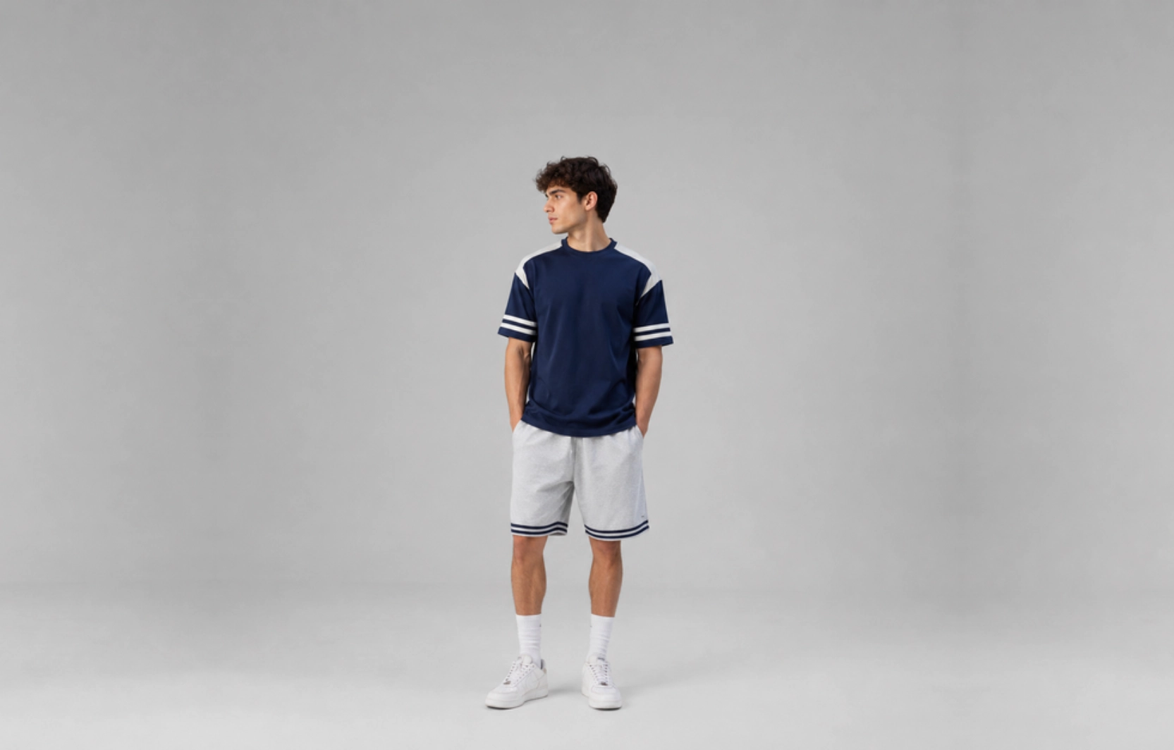 Young man wearing a navy athletic shirt with white stripe accents, light gray shorts, white socks and sneakers, standing against a plain gray studio backdrop.