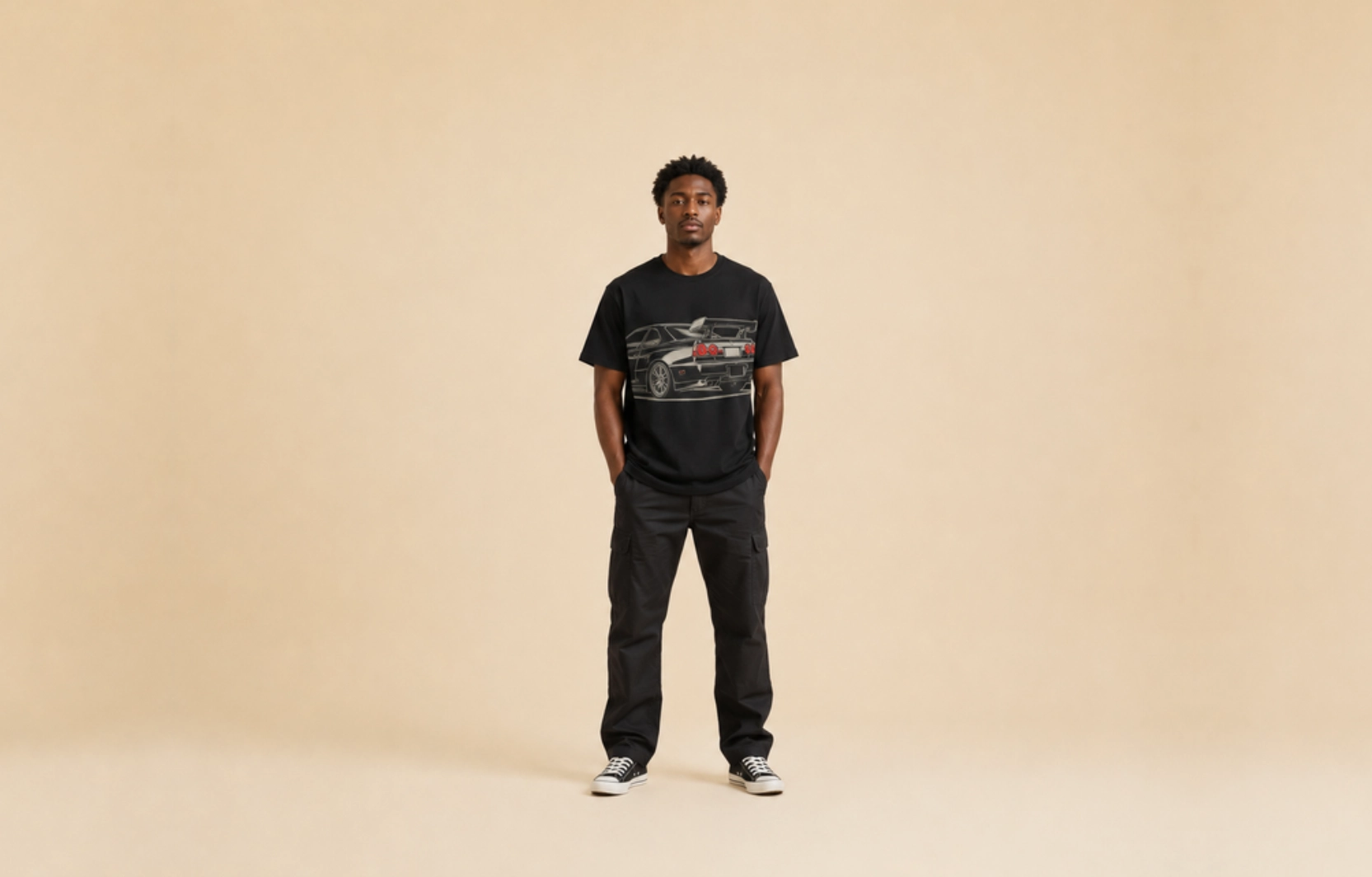 Man wearing a black T-shirt with a car graphic, dark cargo pants, and sneakers stands center against a plain beige background.