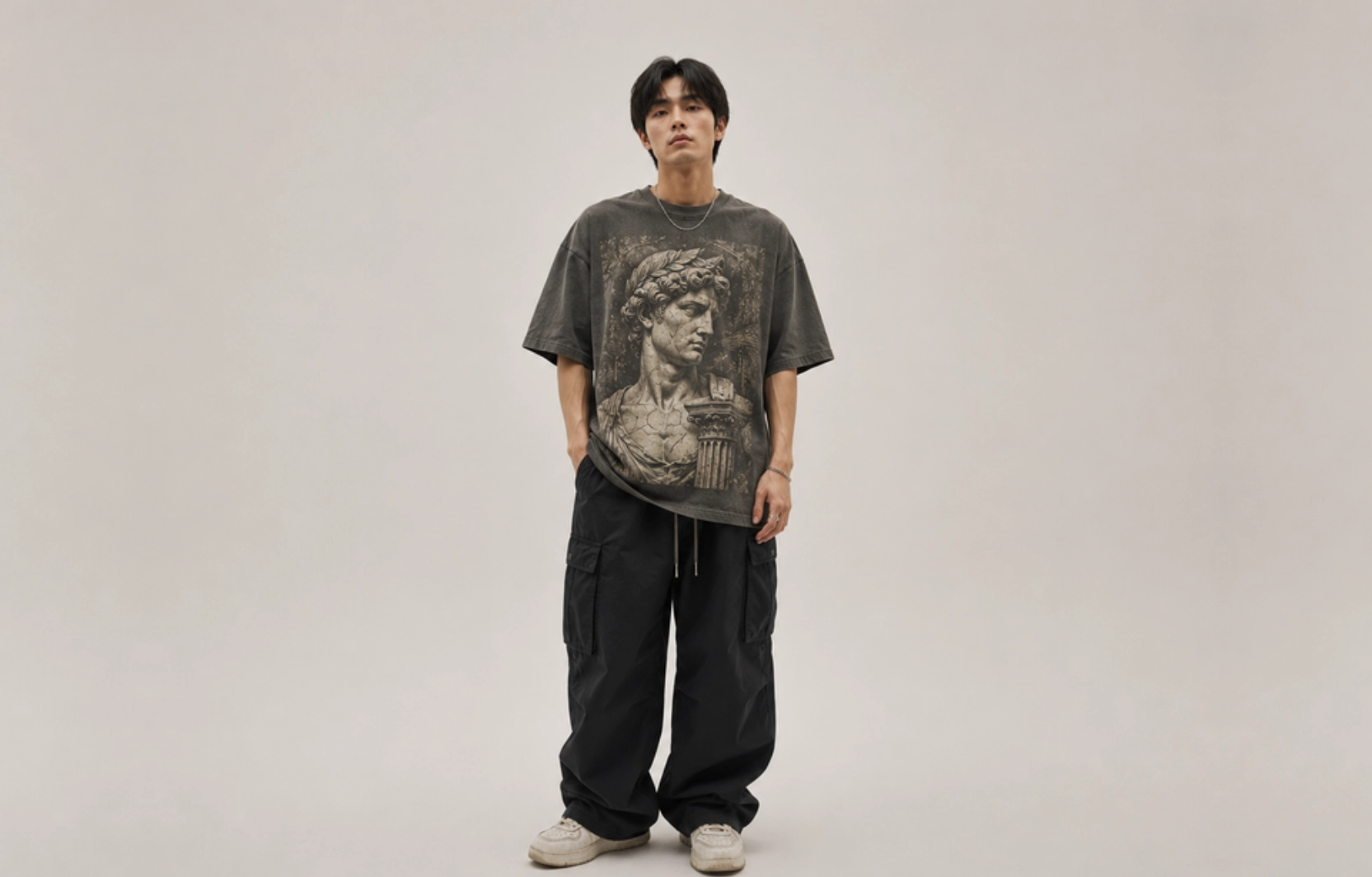 Young man in an oversized dark T-shirt with a classical statue graphic, black cargo pants, and white sneakers, posed against a beige backdrop.