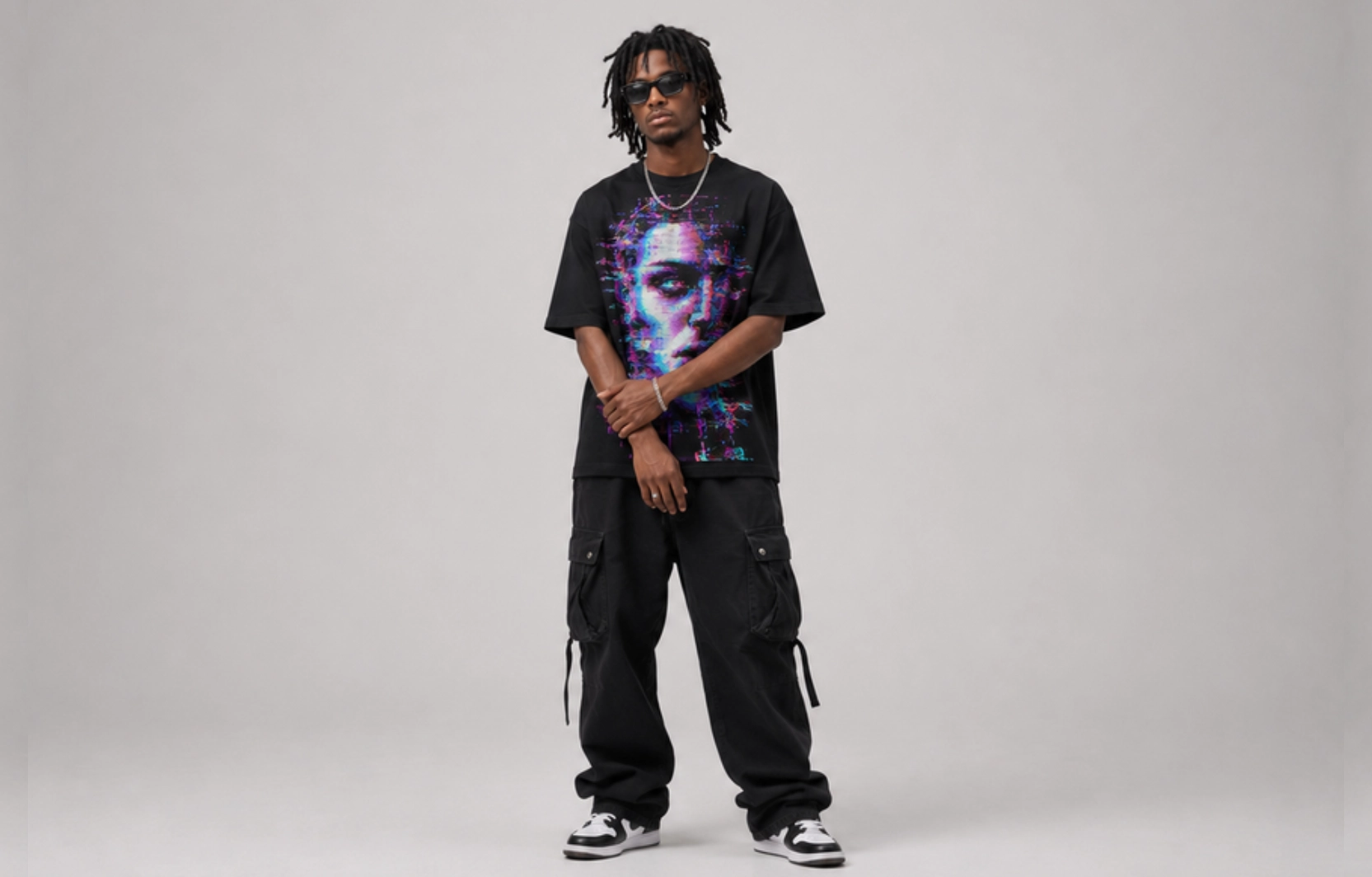 Man wearing a black oversized graphic T-shirt with a neon portrait, black cargo pants, sneakers, and sunglasses, standing against a gray backdrop.