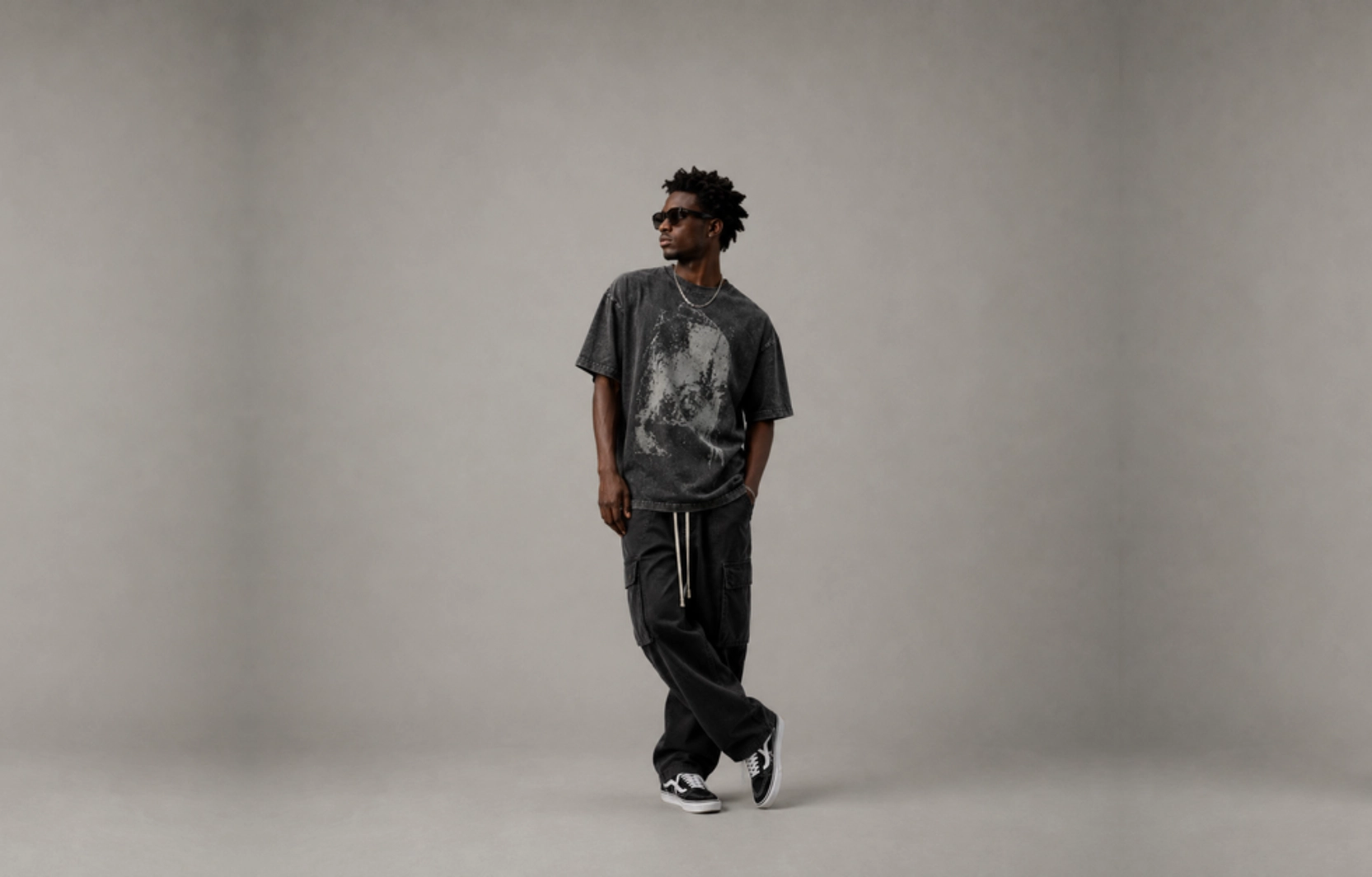 Man wearing a faded black graphic tee, black cargo joggers, and sneakers, posing with one leg crossed in a gray studio setting with sunglasses on.