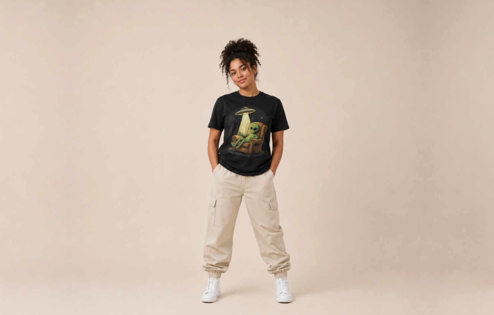 Full-body of a woman in a black graphic tee with an alien in a UFO, beige cargo pants, and white sneakers against a neutral backdrop.