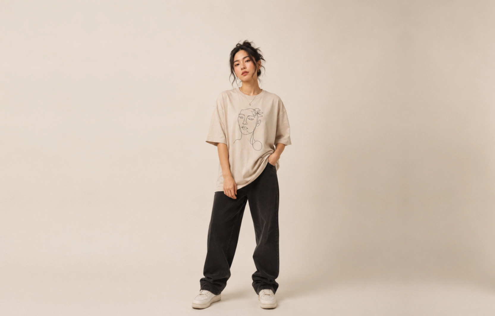 Person standing in a studio wearing a beige oversized T-shirt with a line drawing, black baggy pants, and white sneakers.