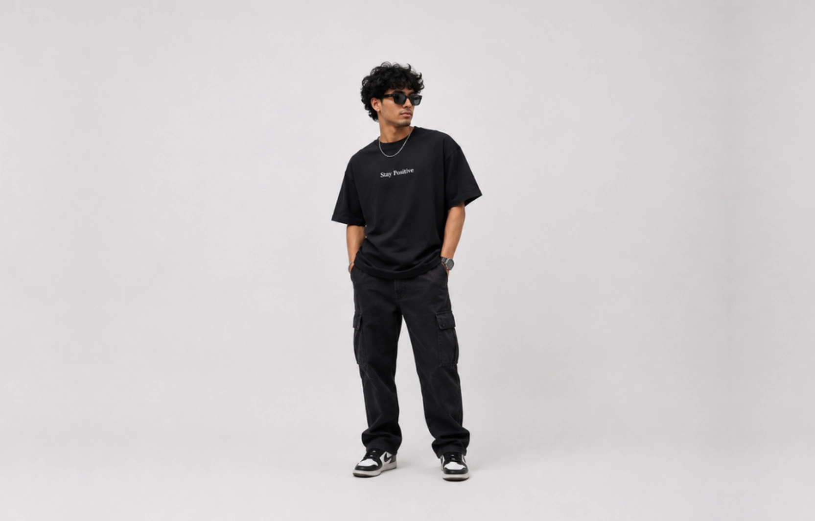 Man in a black oversized T-shirt with 'Stay Positive' slogan, black cargo pants, sneakers, and sunglasses poses against a light gray background.