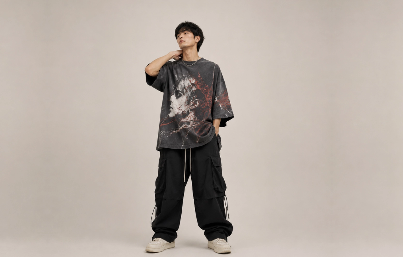 Young man modeling oversized graphic T-shirt with black cargo pants against a neutral studio backdrop, showcasing streetwear style.