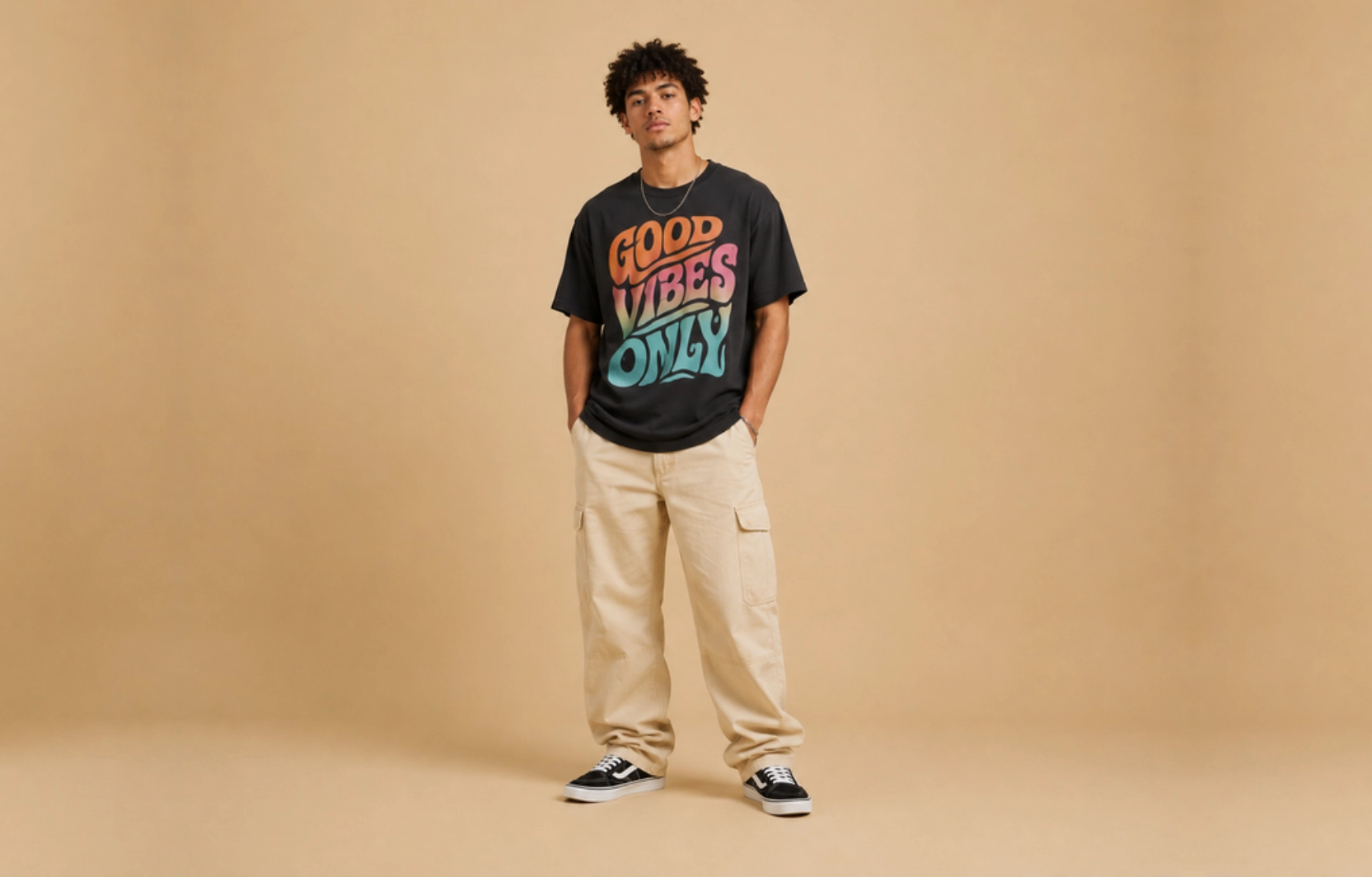 Man standing against a tan backdrop, wearing a black graphic Tโshirt that says 'GOOD VIBES ONLY', beige cargo pants, and black sneakers with white soles.