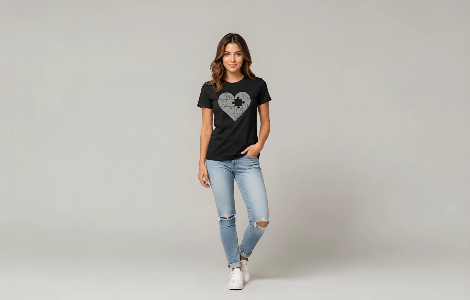 Model wearing a black T-shirt with a heart puzzle graphic, light-wash ripped jeans, and white sneakers in a studio setting.