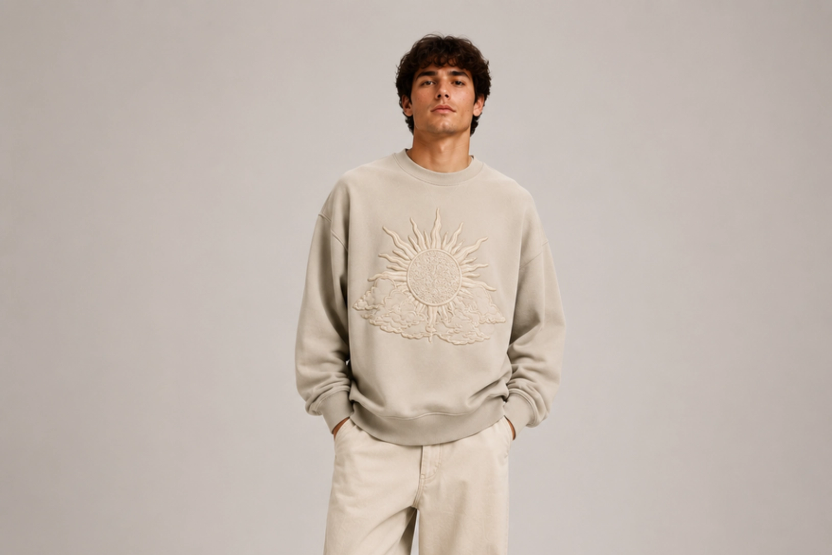 Man wearing a beige sweatshirt with a sunburst emblem embossed on the chest, standing against a neutral backdrop.