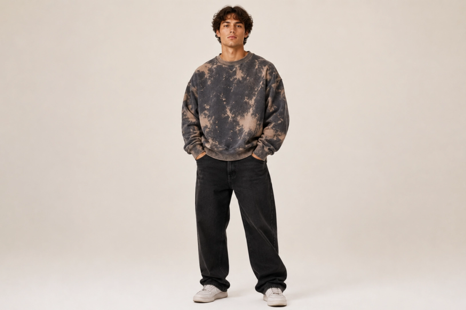 Young man stands facing the camera in a gray‑black tie‑dye sweatshirt, black jeans, and white sneakers, with hands in his pockets.