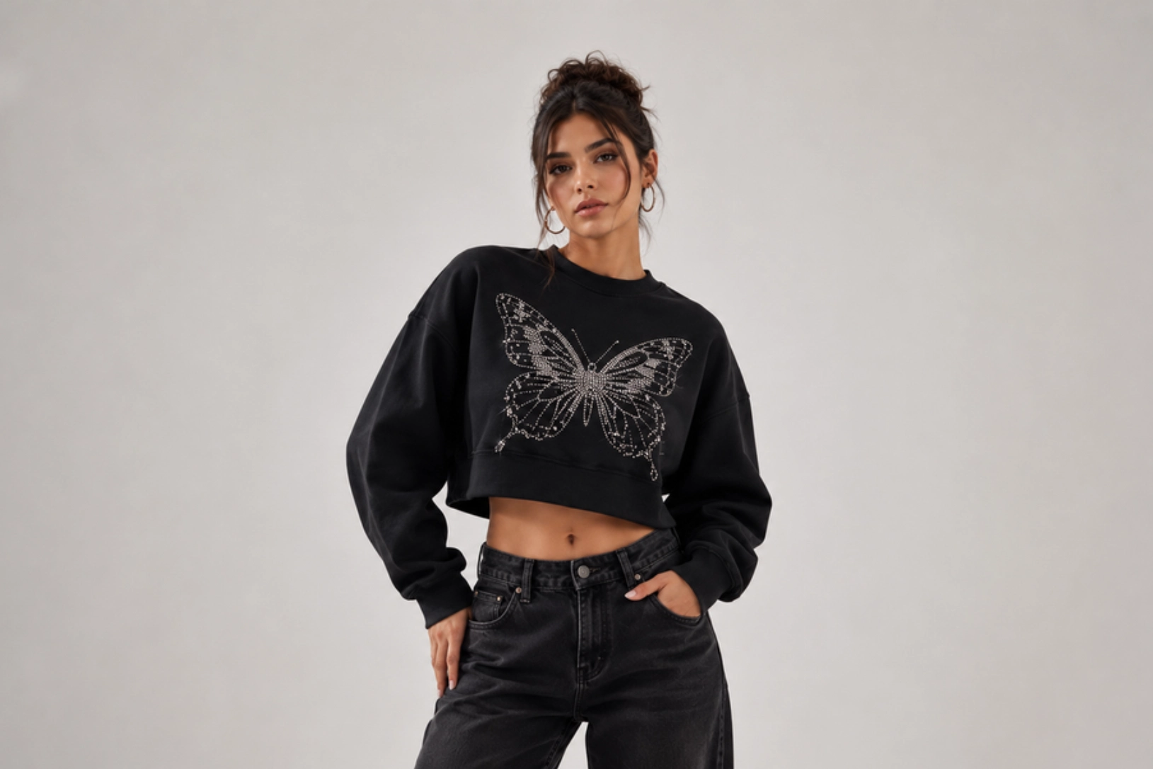 Woman wearing a black cropped sweatshirt with a rhinestone butterfly design, posing with hands in pockets against a light gray backdrop.