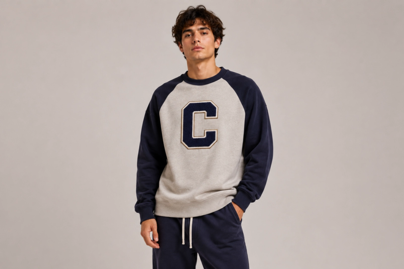 Young man posing in a two-tone sweatshirt (gray body, navy sleeves) with a large navy C logo on the chest.