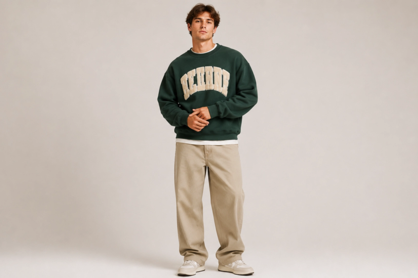 A man in a dark green sweatshirt with beige lettering, beige pants, and white sneakers stands in a neutral studio backdrop with hands loosely clasped.