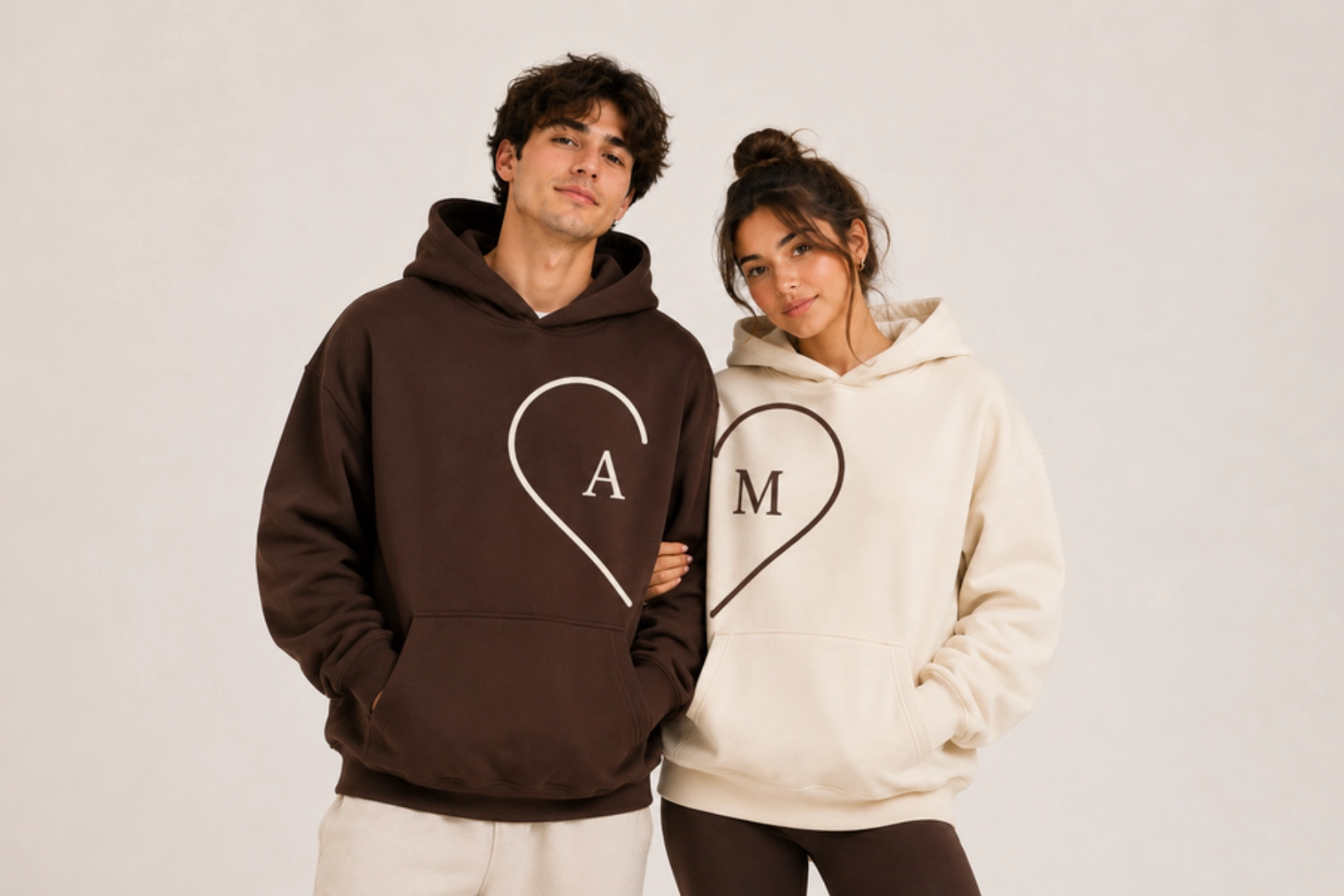 Couple wearing matching oversized hoodies with heart logos: brown hoodie shows letter A, cream hoodie shows letter M, they stand together smiling.