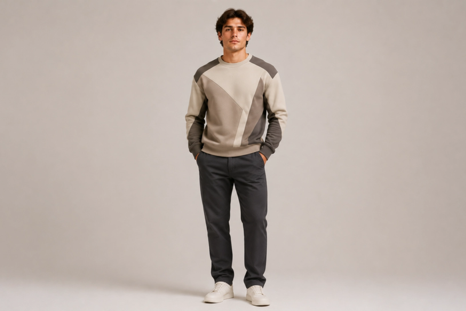 Man standing against a neutral backdrop in a beige and gray geometric color-block sweater, dark pants, and white sneakers.