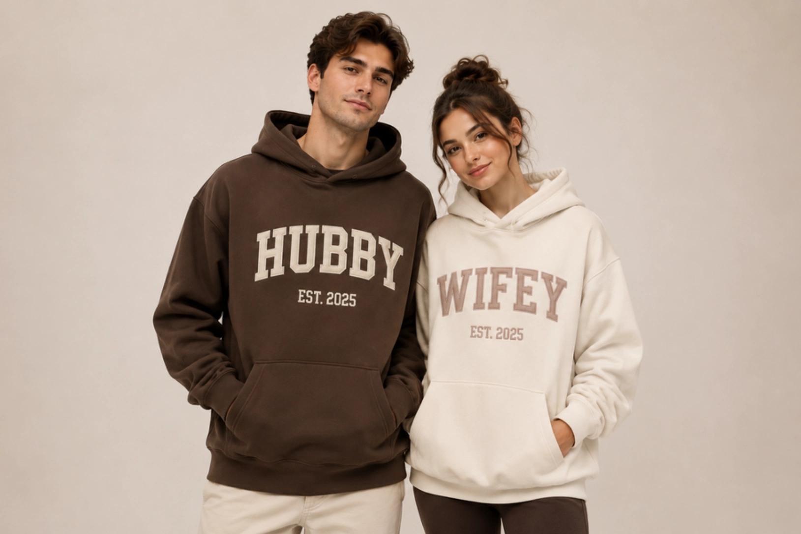 Couple wearing matching hoodies: man in brown hoodie that says 'HUBBY EST. 2025' and woman in cream hoodie that says 'WIFEY EST. 2025' against a neutral backdrop.