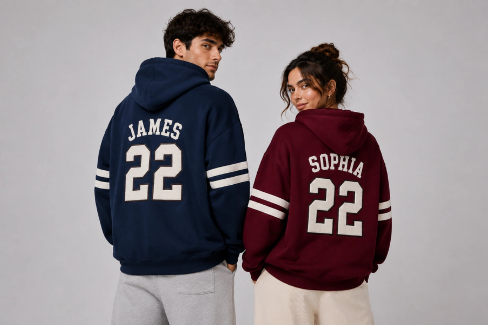 Two people with backs to the camera wearing oversized hoodies: navy hoodie shows JAMES 22, maroon hoodie shows SOPHIA 22 with white stripes on sleeves