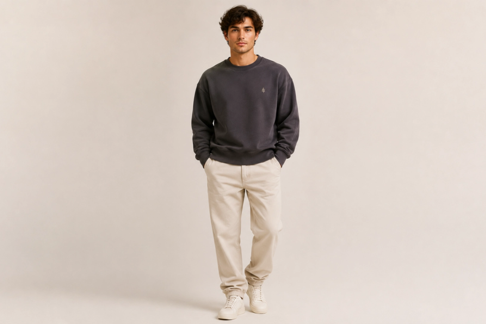 Man in a dark gray sweatshirt, beige pants, and white sneakers stands with hands in pockets against a neutral studio backdrop.