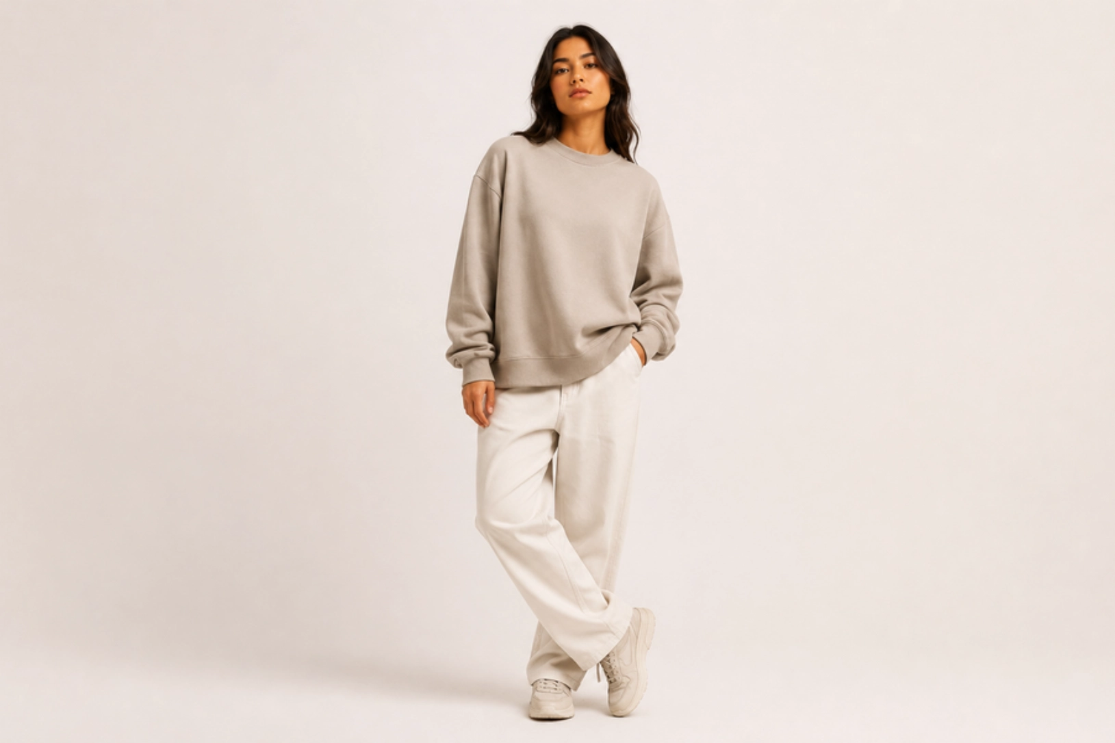Woman in an oversized beige sweatshirt and light pants stands against a pale backdrop, hands in pockets.