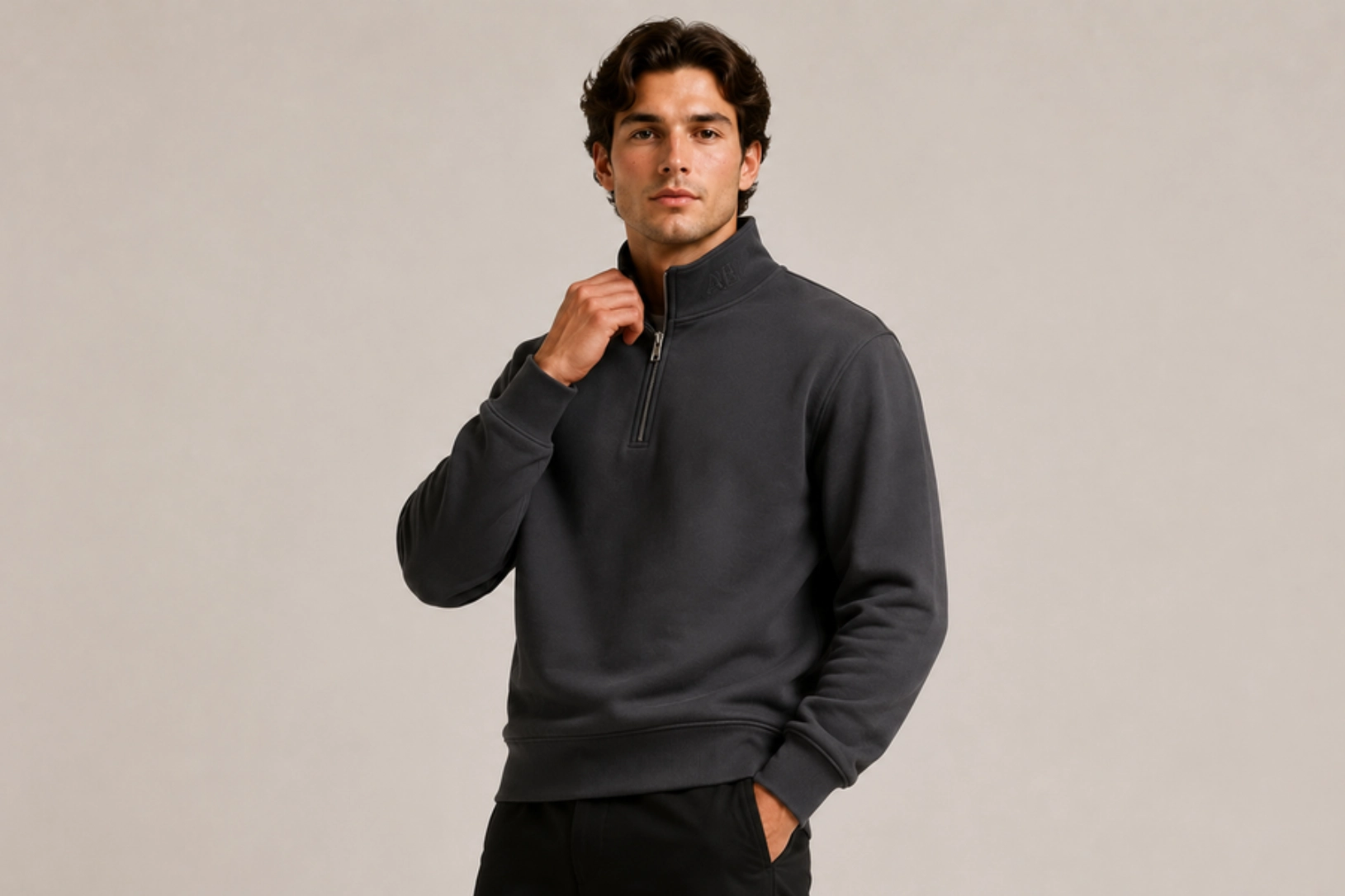 Young man in a dark gray quarter-zip fleece pullover, touching the collar against a beige background.