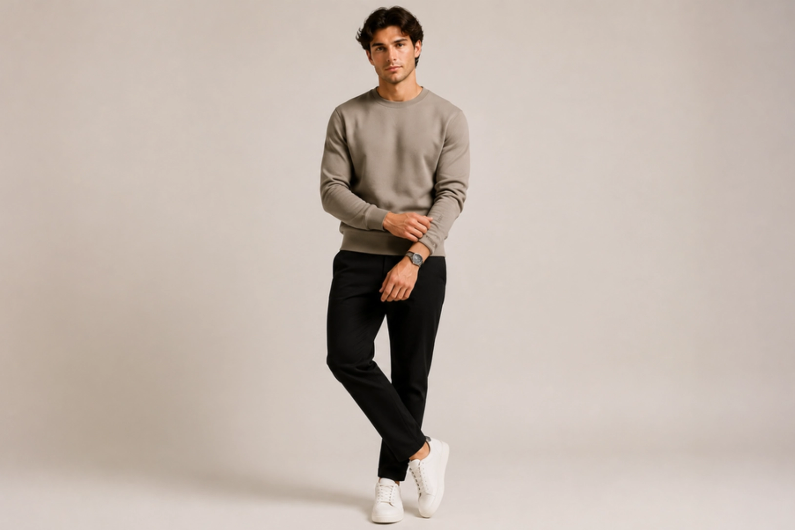 Man wearing a beige crewneck sweater, black pants, and white sneakers posing with one leg crossed against a neutral backdrop.