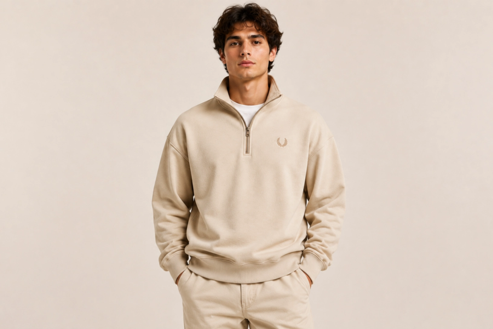 Young man wearing a beige fleece pullover with a half-zip and matching joggers, hands in pockets, neutral backdrop.