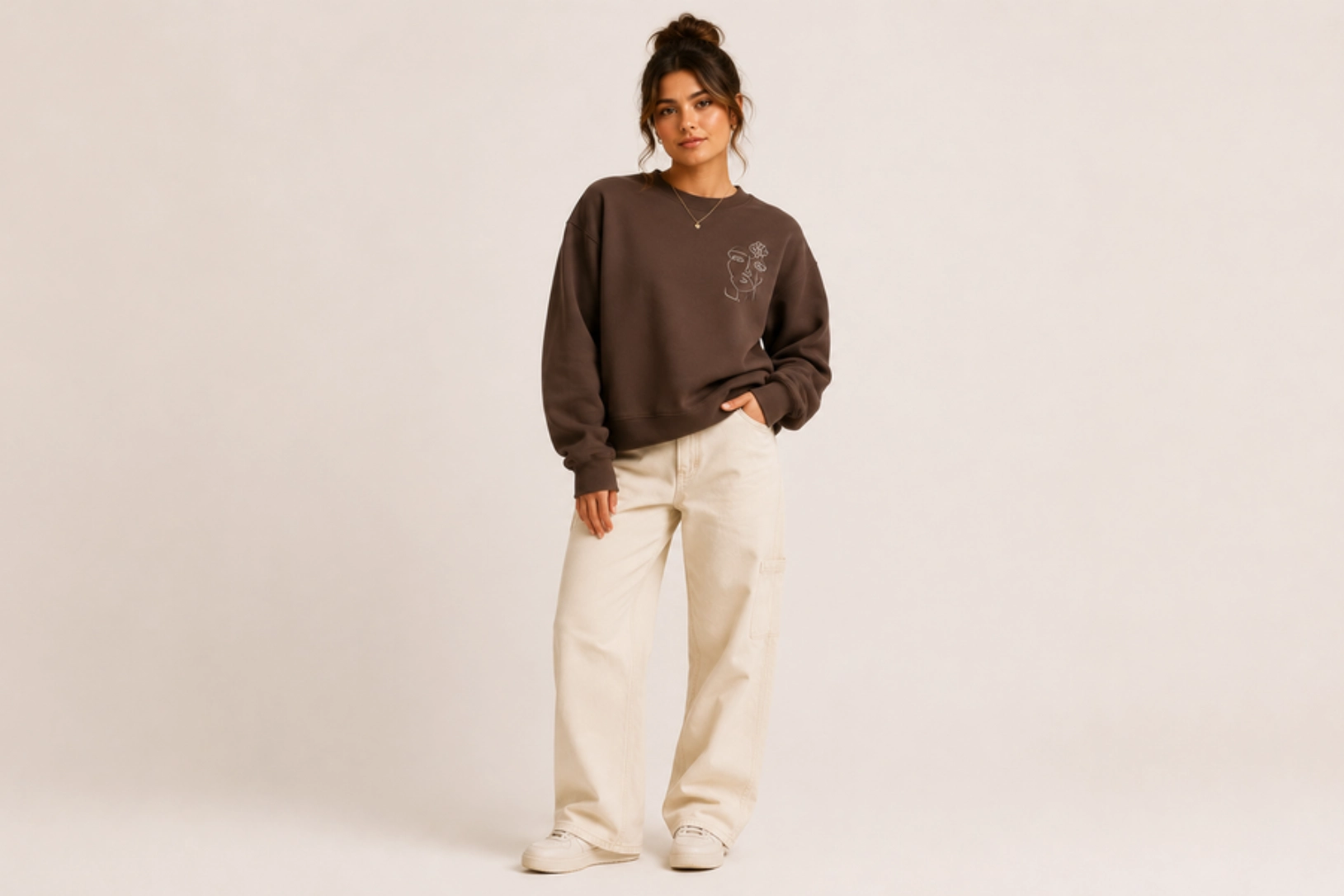 Woman wearing a brown oversized sweatshirt with a small chest illustration, beige cargo pants, and white sneakers in a studio setting.