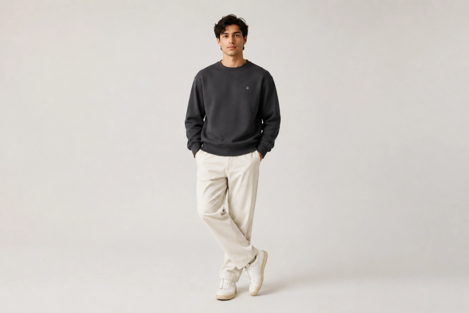 Young man in a dark gray sweatshirt, beige chinos, and white sneakers stands casually with hands in pockets against a light neutral backdrop.