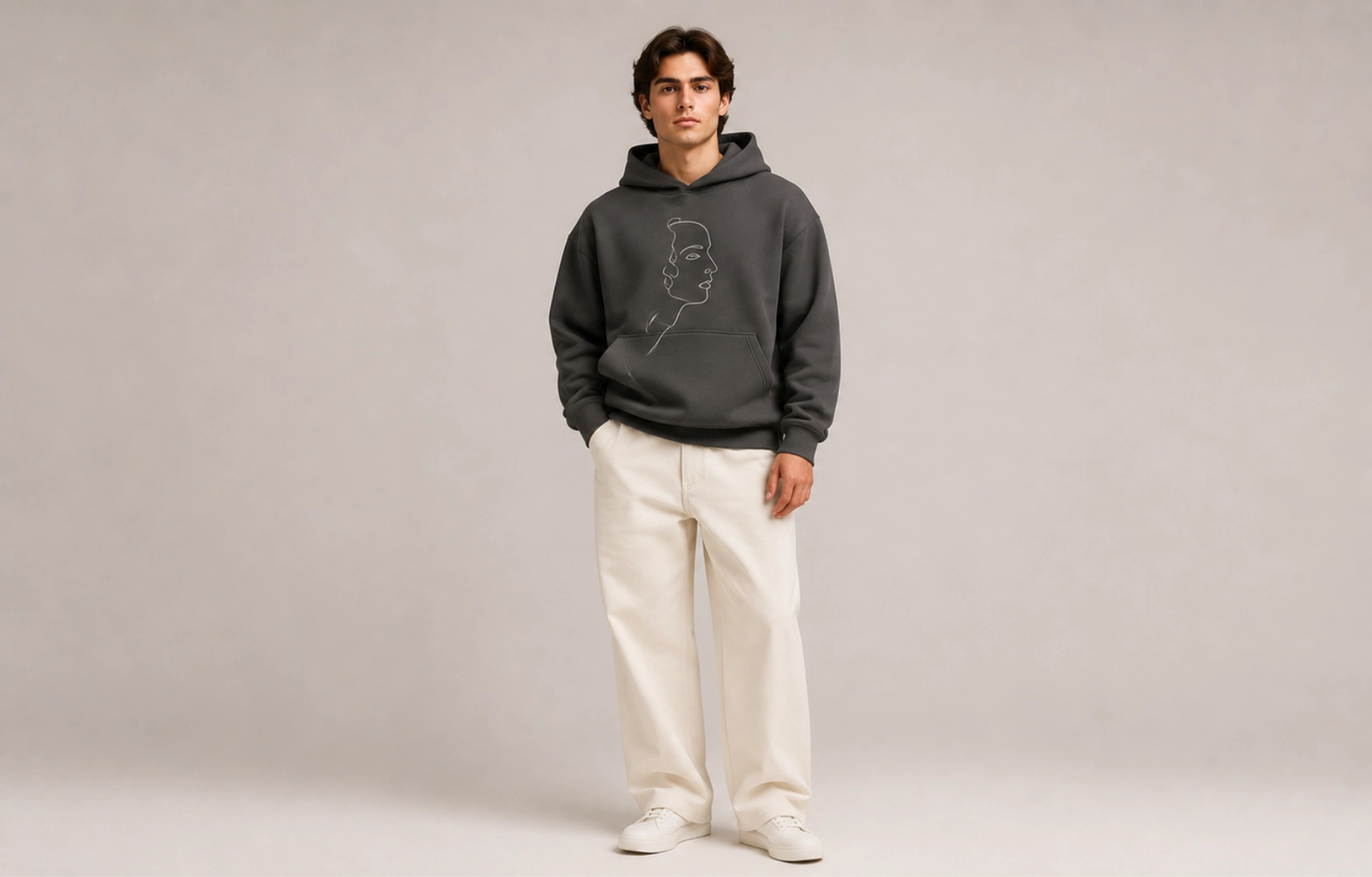 Young man wearing a dark gray hoodie with a subtle line-art portrait, paired with beige wide-leg pants and white sneakers in a neutral studio setting.