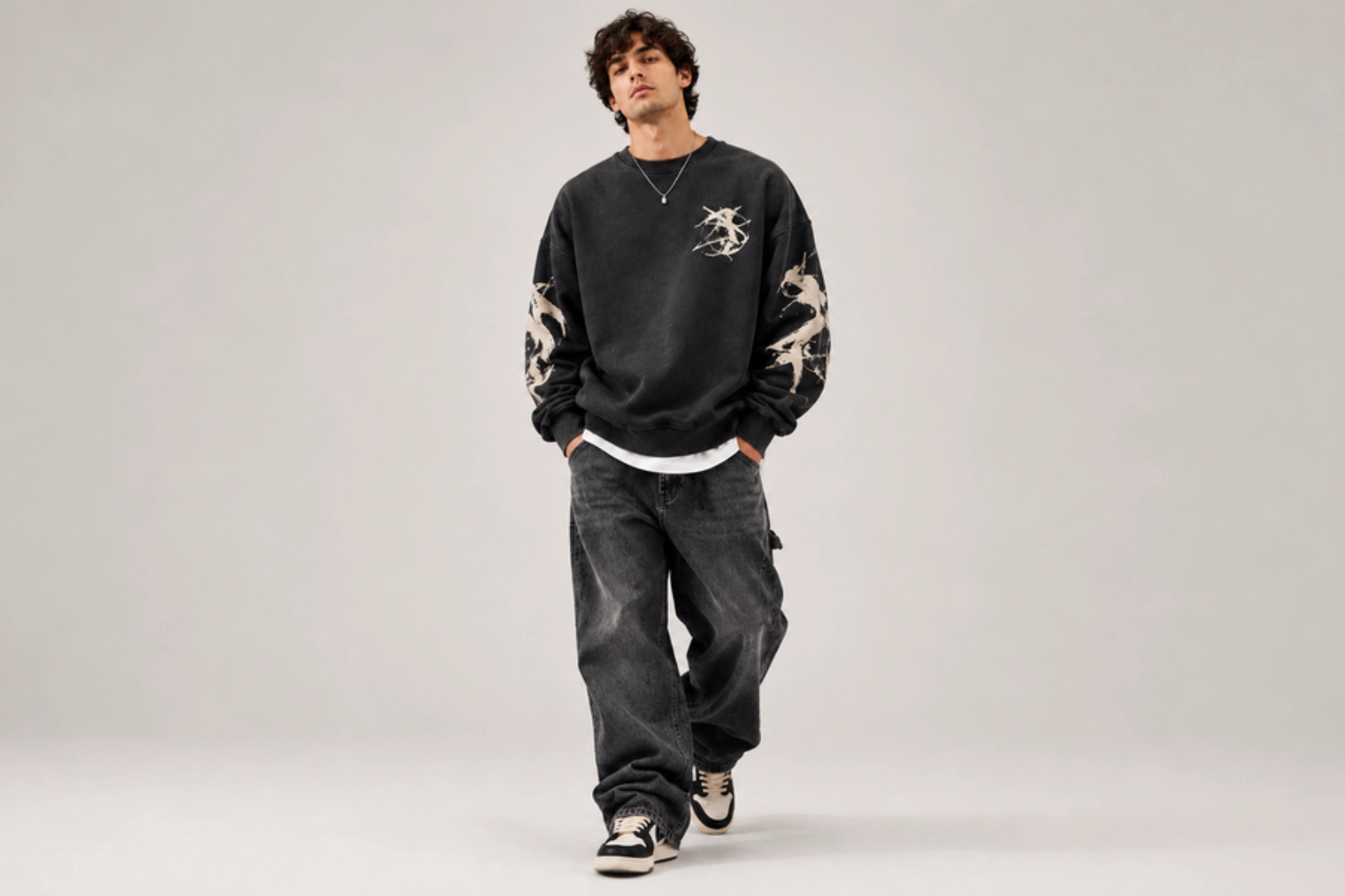 Model wearing an oversized black sweatshirt with bleach splatters, layered over a white shirt, and loose dark jeans with sneakers in a studio.
