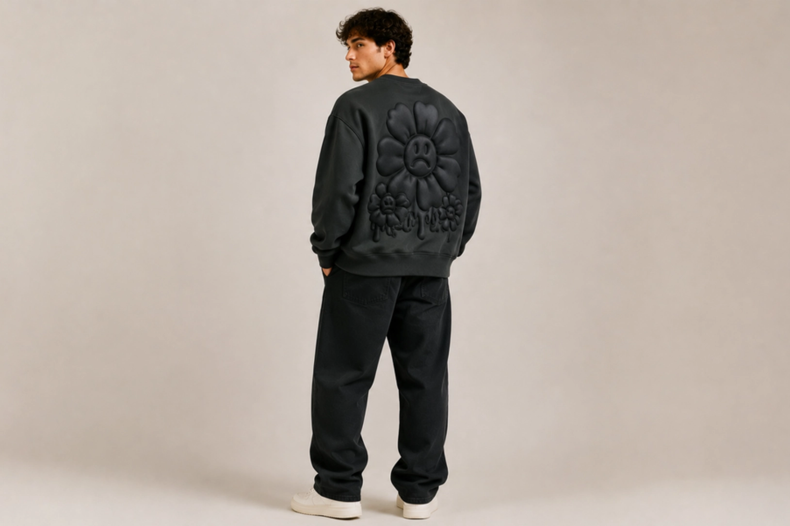 Person seen from behind wearing a dark oversized sweatshirt with large embossed flower design on the back, black pants, white sneakers.