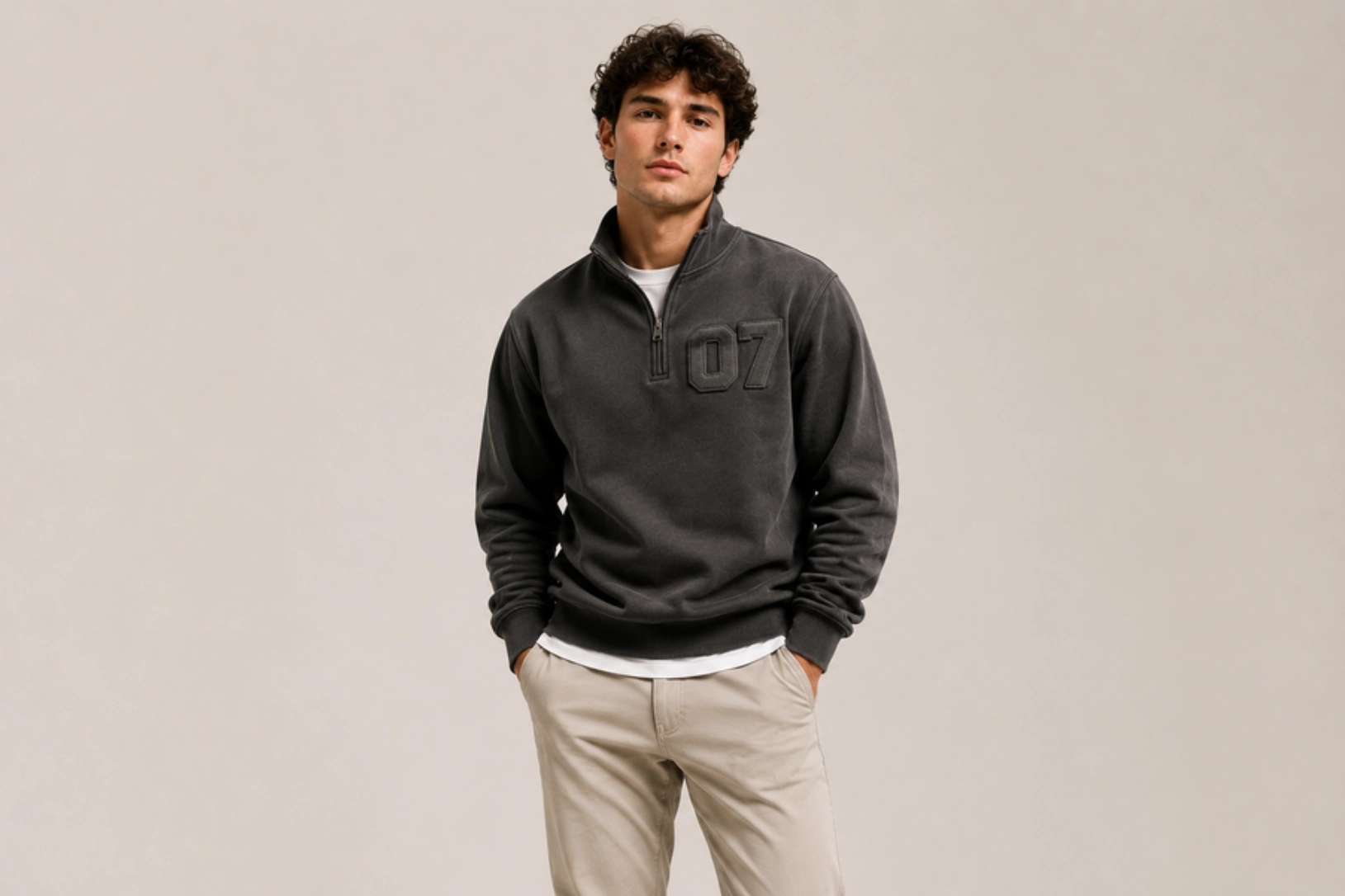 Young man wearing a dark gray pullover with '107' on the chest, white undershirt, beige pants, hands in pockets against a neutral backdrop.