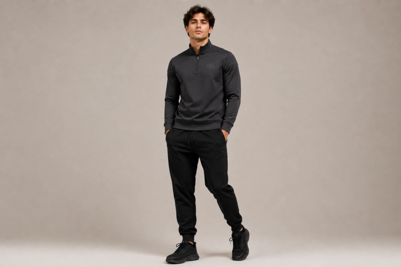 Man wearing a dark, long-sleeve half-zip athletic top and black joggers, standing with hands in pockets against a neutral backdrop, full body.