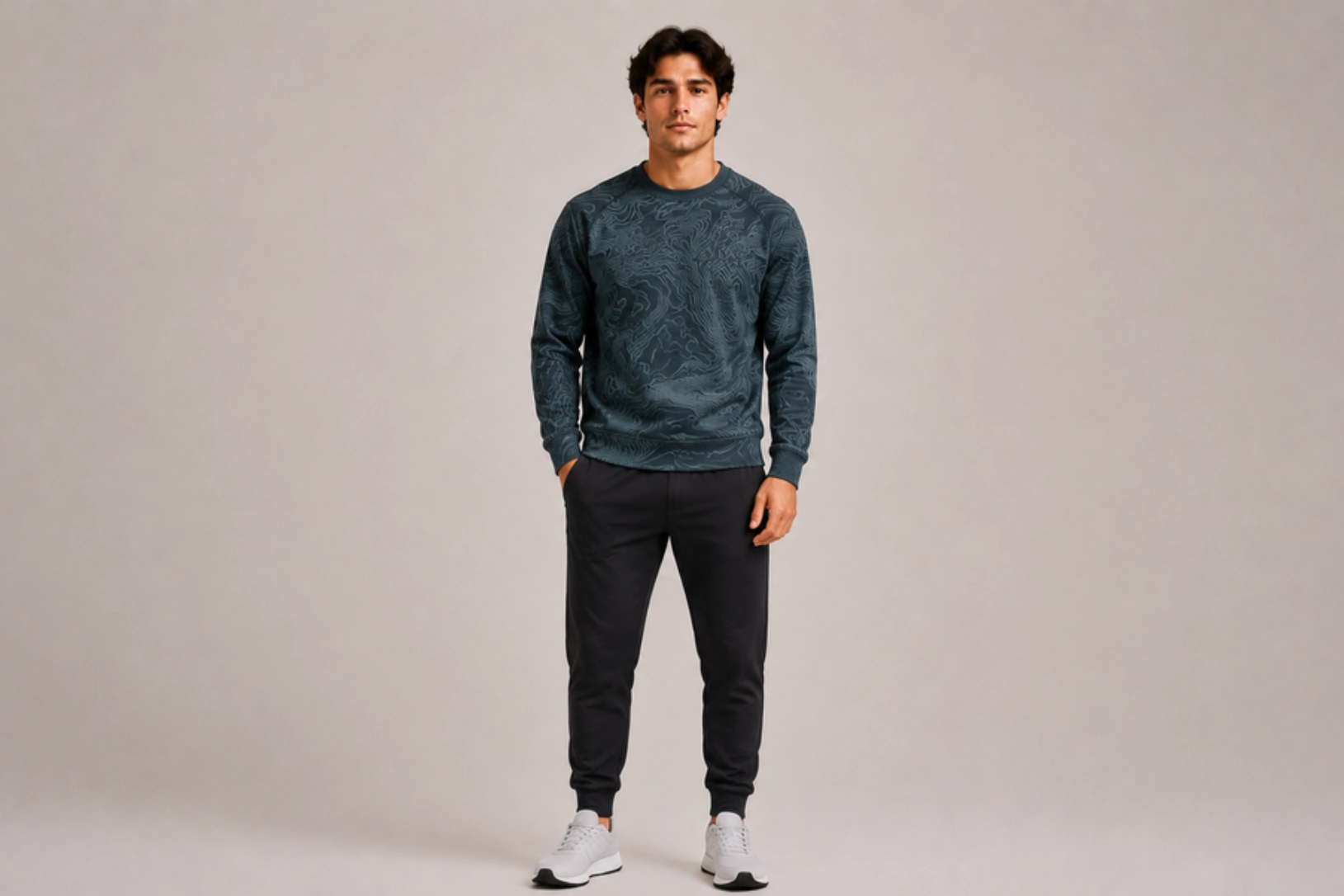 Man standing in a neutral studio, wearing a teal patterned crewneck sweatshirt, black joggers, and white sneakers.