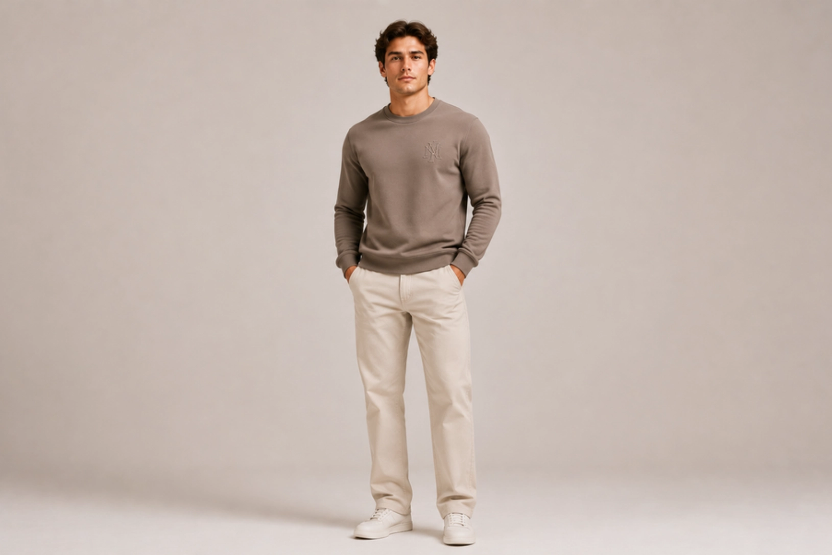 Young man in a taupe crewneck sweatshirt and beige pants stands with hands in pockets against a neutral studio backdrop, wearing white sneakers.