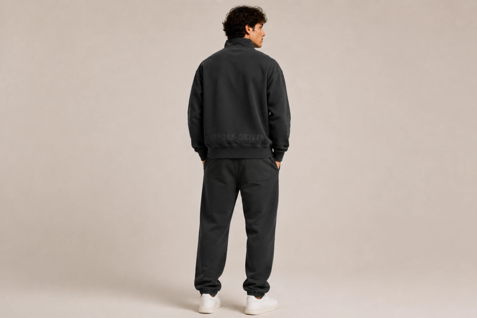 Person standing with back to camera, wearing a dark tracksuit and white sneakers in a neutral studio setting.