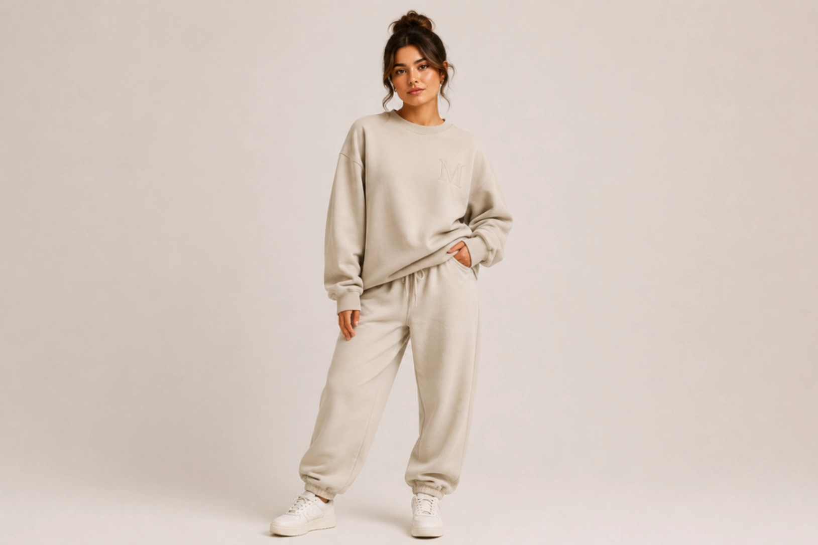 Woman modeling a beige oversized sweatshirt and matching jogger pants with white sneakers in a neutral studio backdrop.