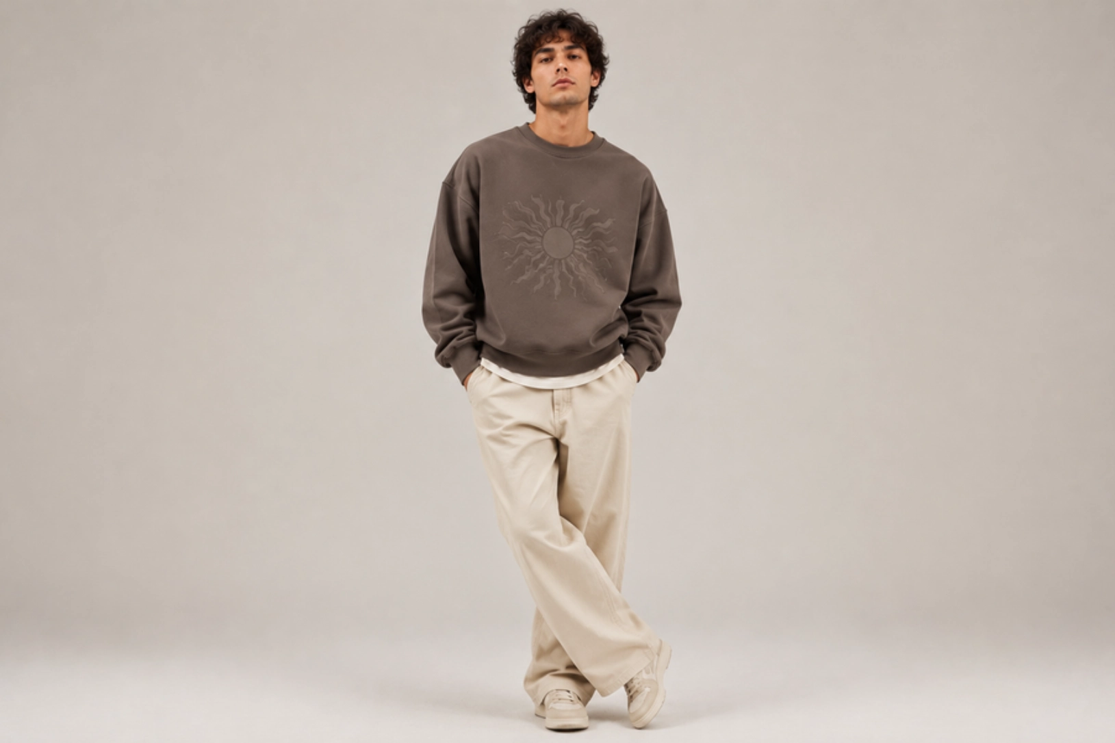 Young man in a brown sun-graphic sweatshirt and beige pants posing with hands in pockets in a neutral studio setting.