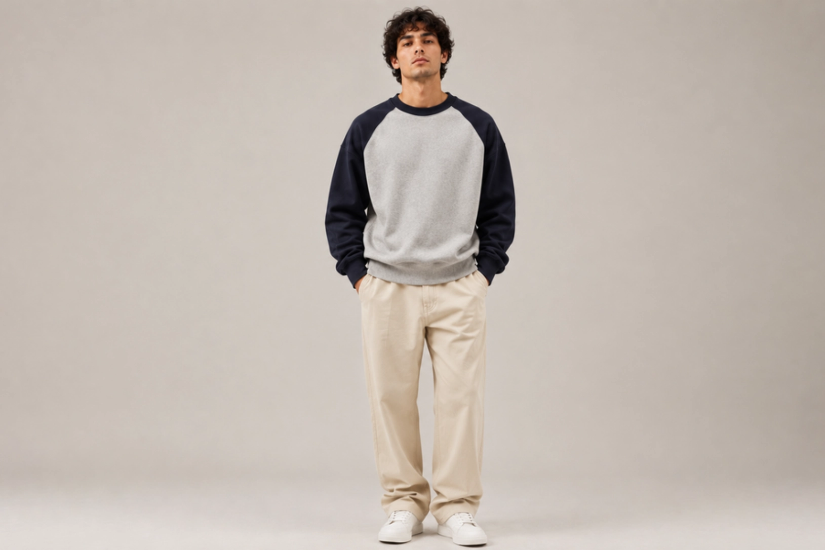 Man standing in a neutral studio wearing a gray body with navy raglan sleeves sweatshirt, beige pants, and white sneakers, hands in pockets.
