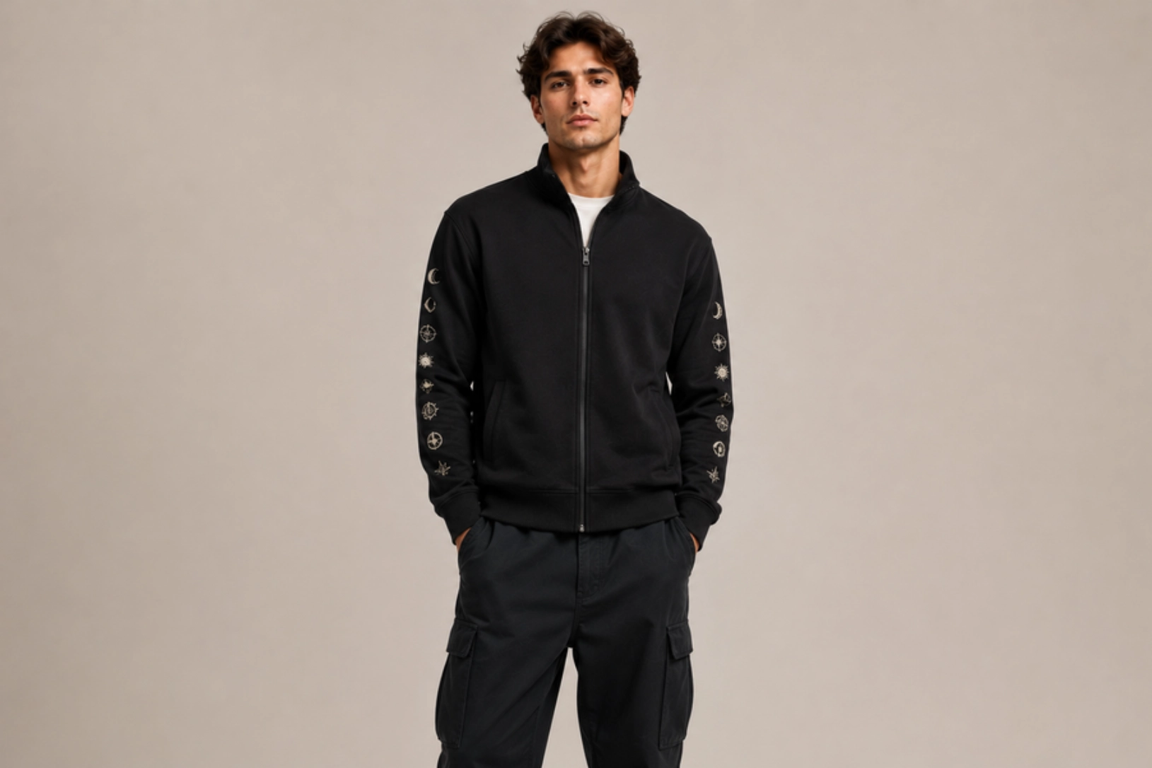 Young man in a black zip-up jacket with embroidered symbols running down the sleeves, standing against a beige studio backdrop with hands in pockets.