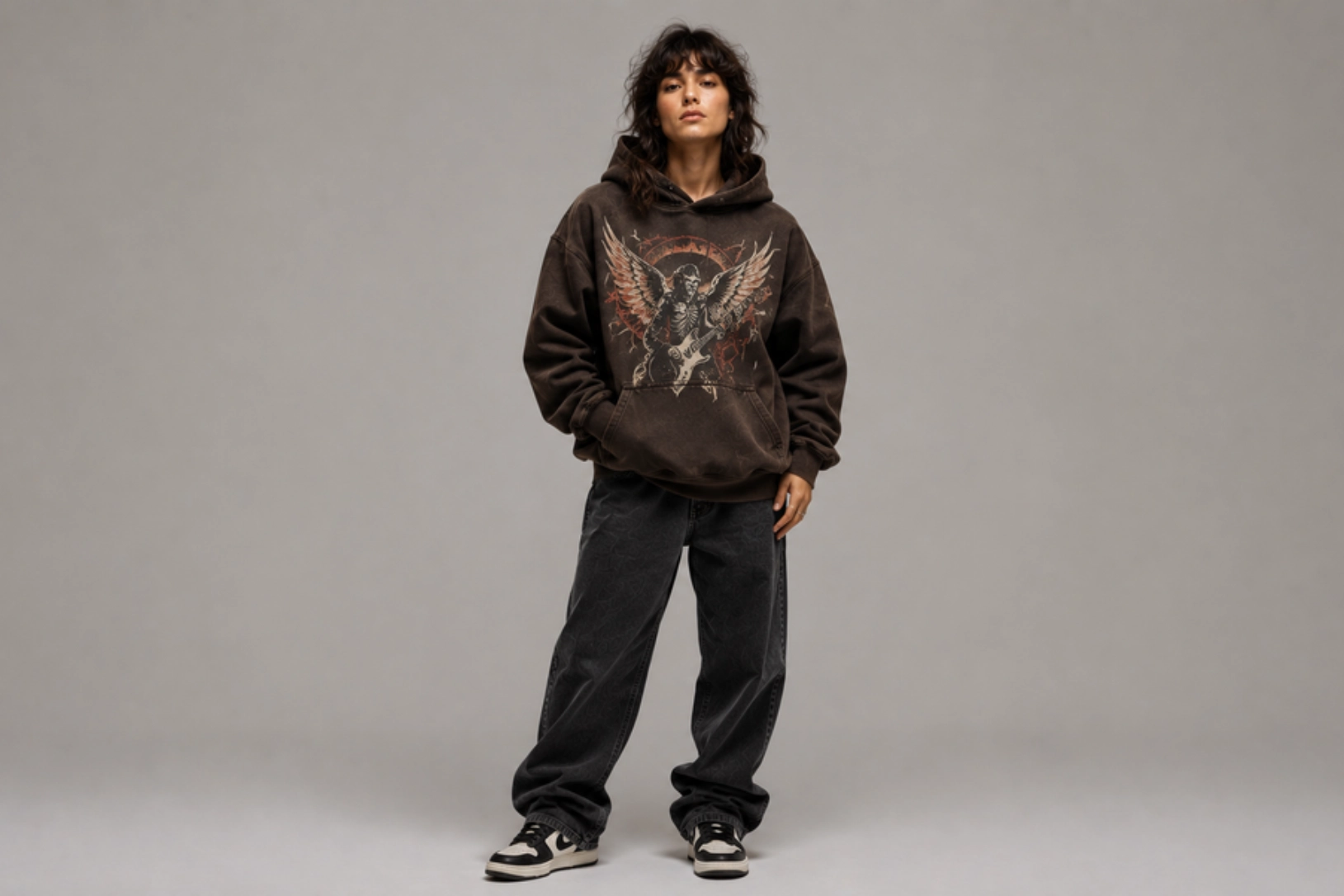 Person standing in a brown oversized hoodie with a graphic of an angel and wings, dark baggy jeans, and sneakers, posing casually against a neutral backdrop.