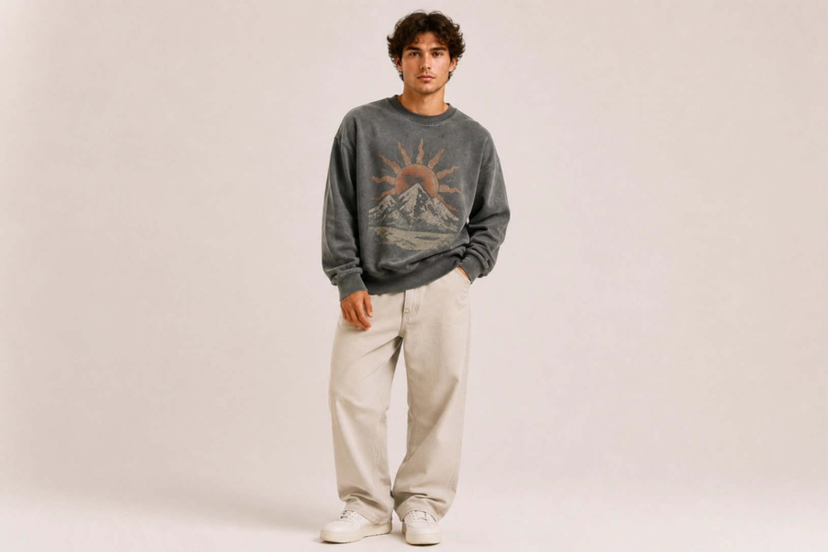 Young man stands in a neutral studio wearing a faded gray graphic sweatshirt with mountains and a sun, beige baggy pants, and white sneakers.