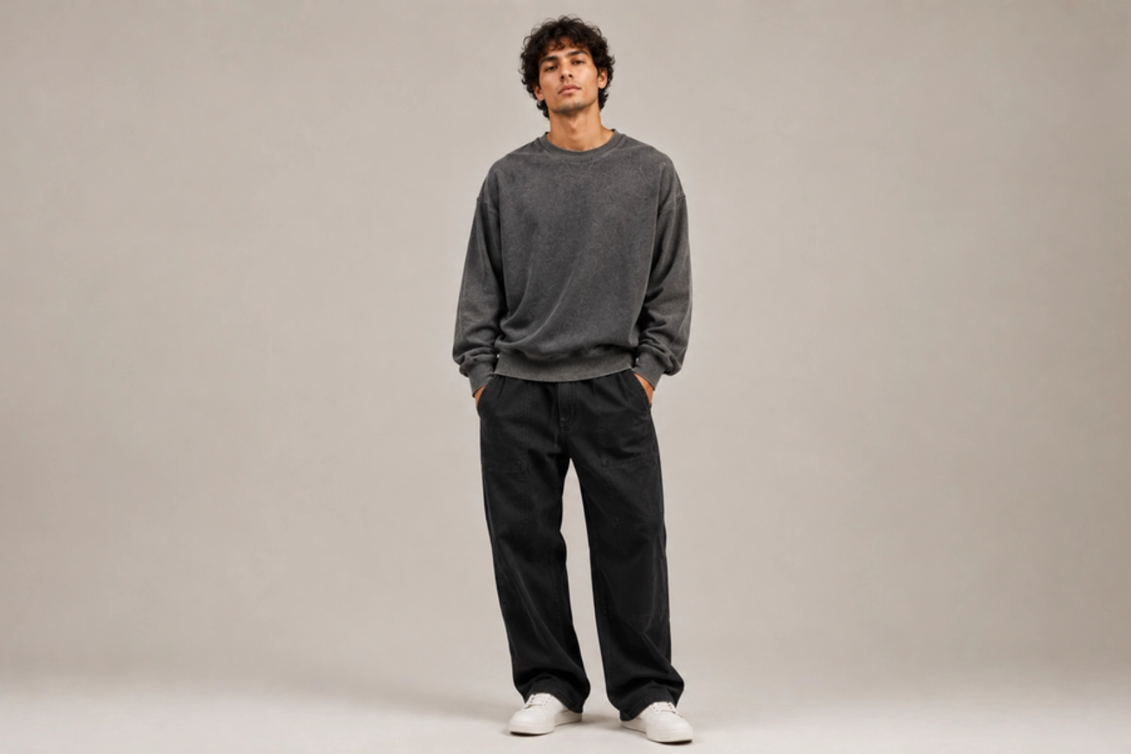 Full-body shot of a man standing in a gray oversized sweatshirt, black pants, and white sneakers against a neutral backdrop.