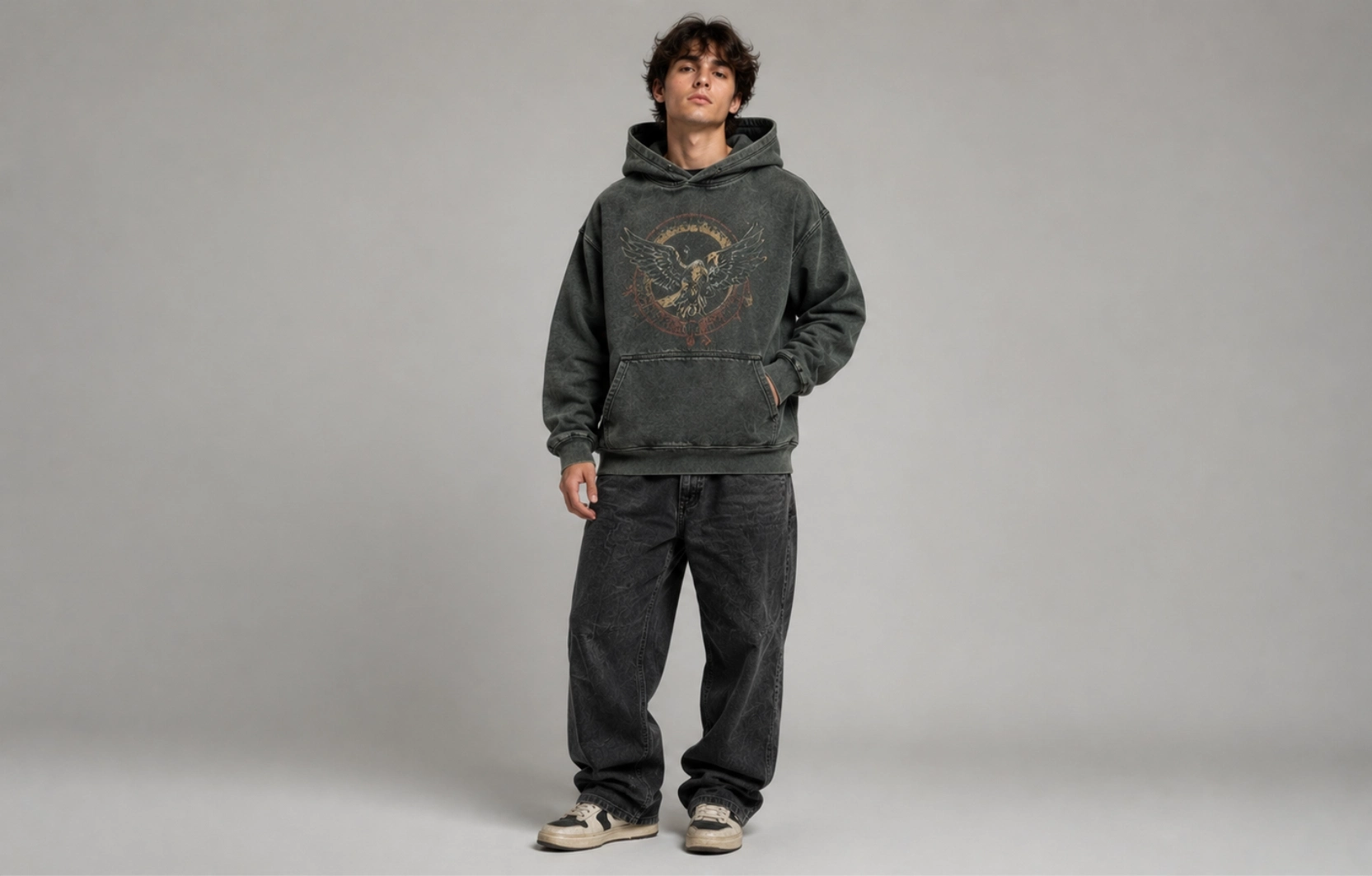 Young man stands in a gray studio wearing a faded charcoal hoodie with a winged emblem, black jeans, and sneakers.