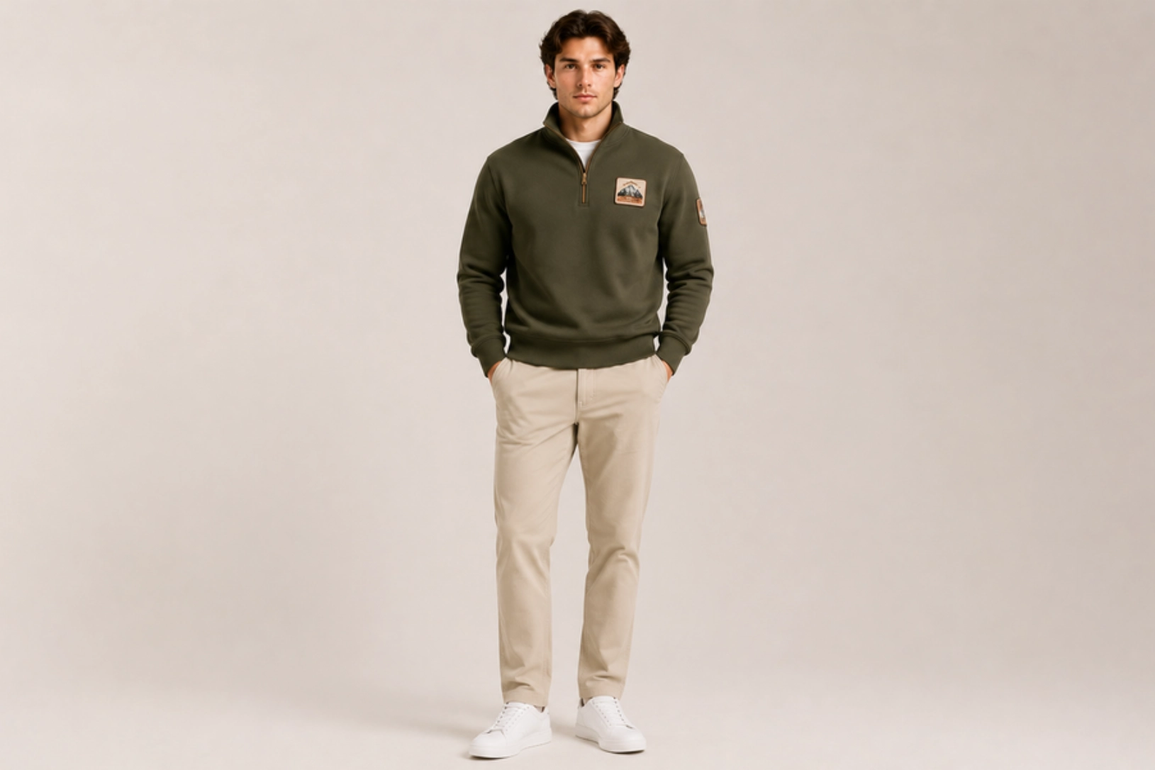 Young man in a studio wearing an olive green quarter-zip sweater with patches, beige pants, and white sneakers.