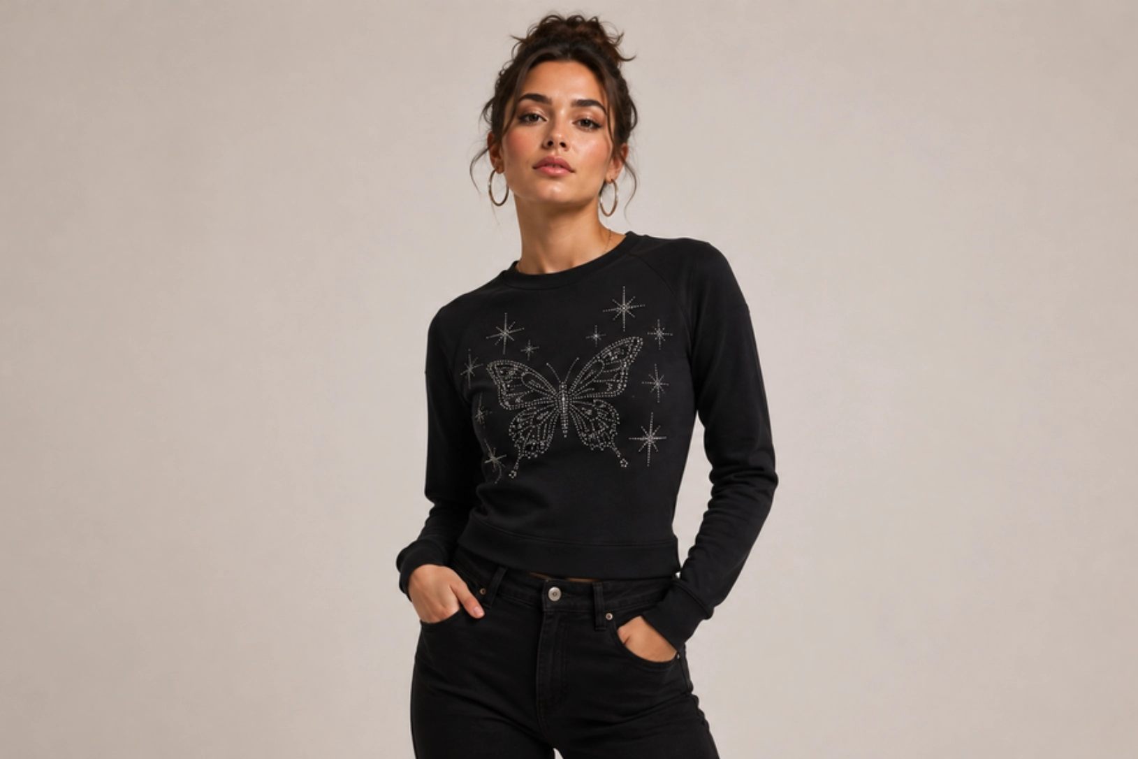 Woman posing in a black sweatshirt featuring a rhinestone butterfly and star pattern, hands in pockets against a neutral backdrop.