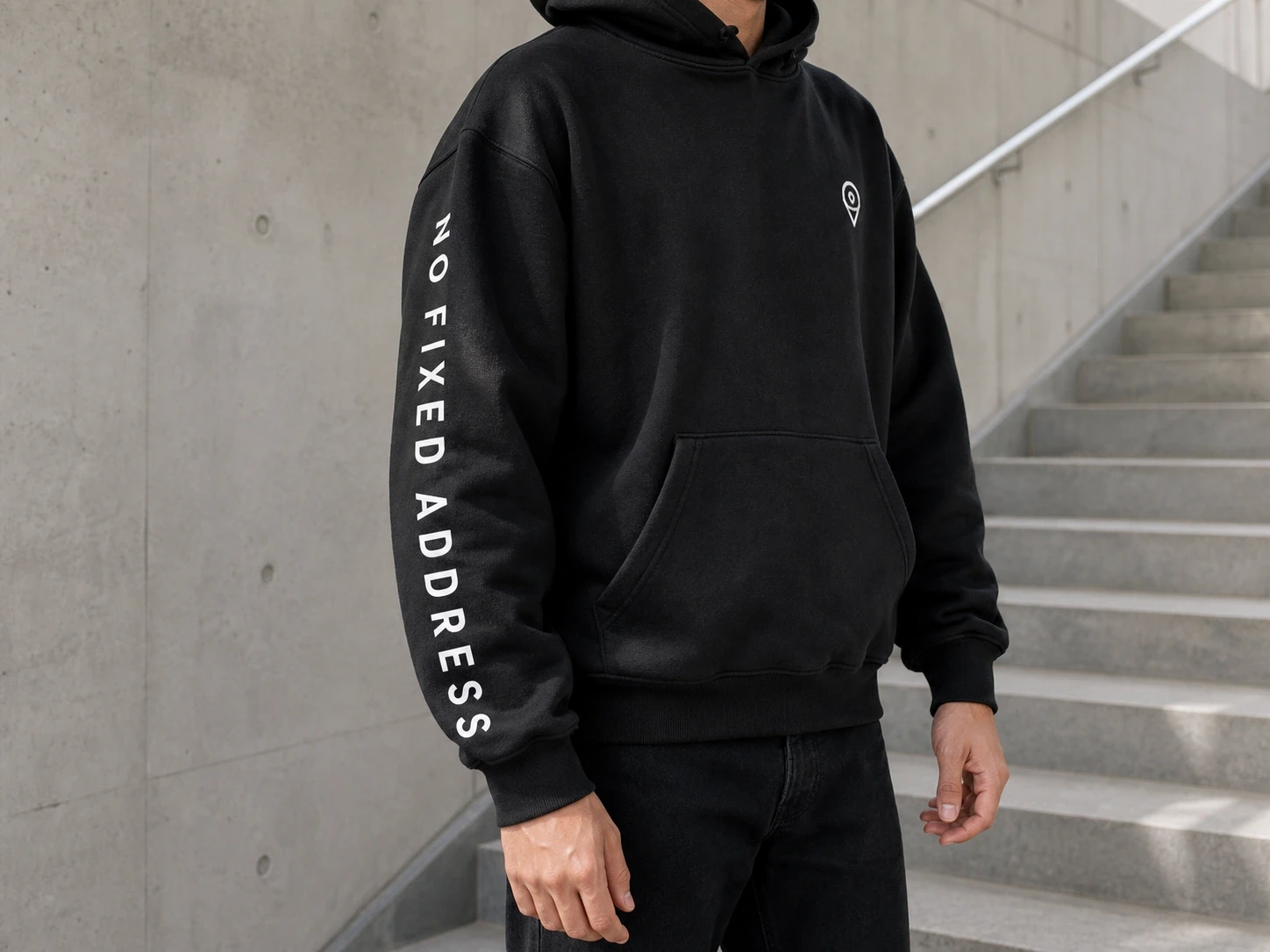 Person in black hoodie with "NO FIXED ADDRESS" on sleeve, standing by concrete steps. Minimalist, urban style.