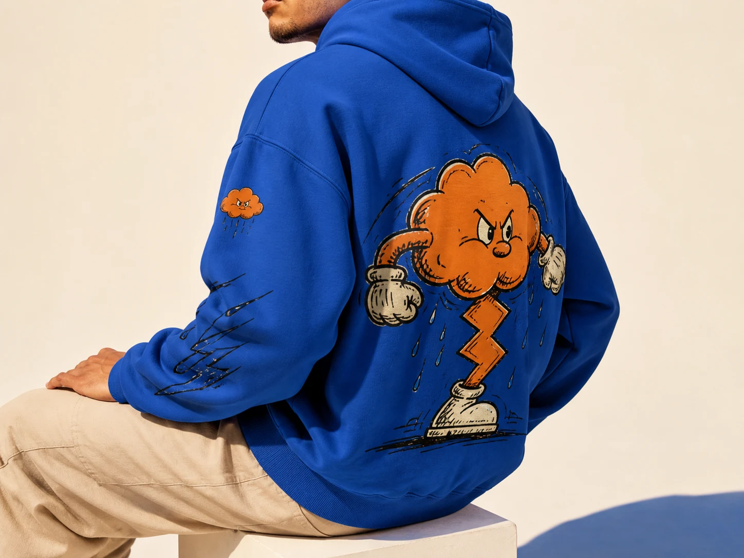 Man in blue hoodie with cartoon cloud and lightning design sits on a white block, wearing beige pants against a neutral background.