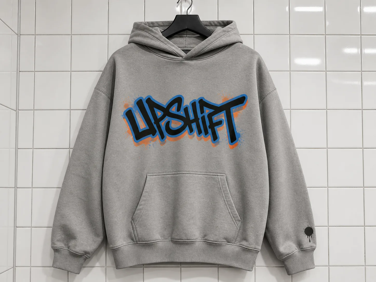 Gray hoodie with "UPSHIFT" in bold graffiti font, blue and orange accents, hangs on a tiled wall.